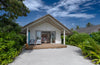 Bungalow with a wooden deck surrounded by greenery under a blue sky.