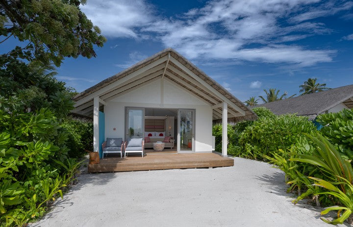 Bungalow with a wooden deck surrounded by greenery under a blue sky.