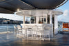 Modern outdoor bar area with white chairs and tables on a deck with a cityscape in the background.