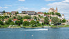 Town on a hill overlooking a river with boats, under a blue sky with clouds.