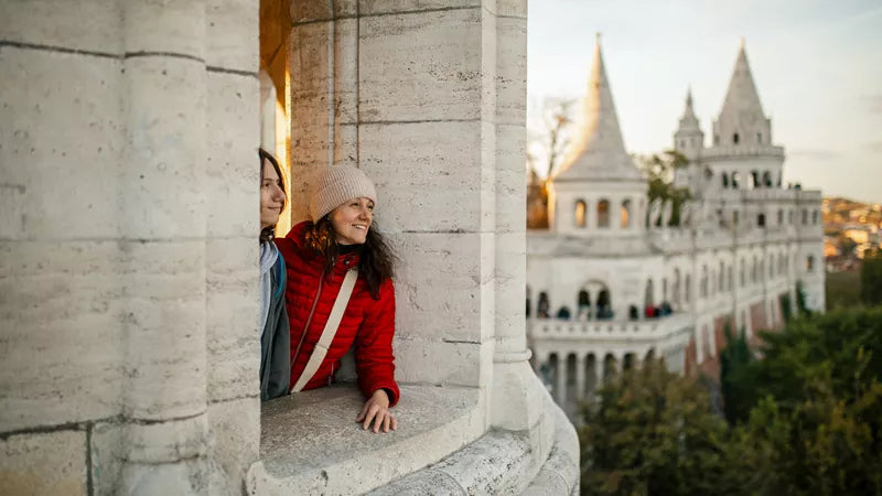 Two people looking out from a stone balcony with a scenic view of a castle-like building.