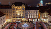 Nighttime view of a square with illuminated buildings and people gathered around a central fountain.