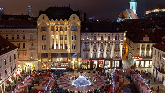 Nighttime view of a square with illuminated buildings and people gathered around a central fountain.