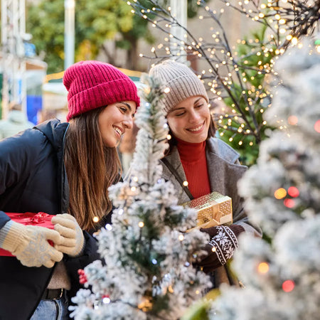 Two women exchanging Christmas presents in a festive outdoor setting with lights and decorations.